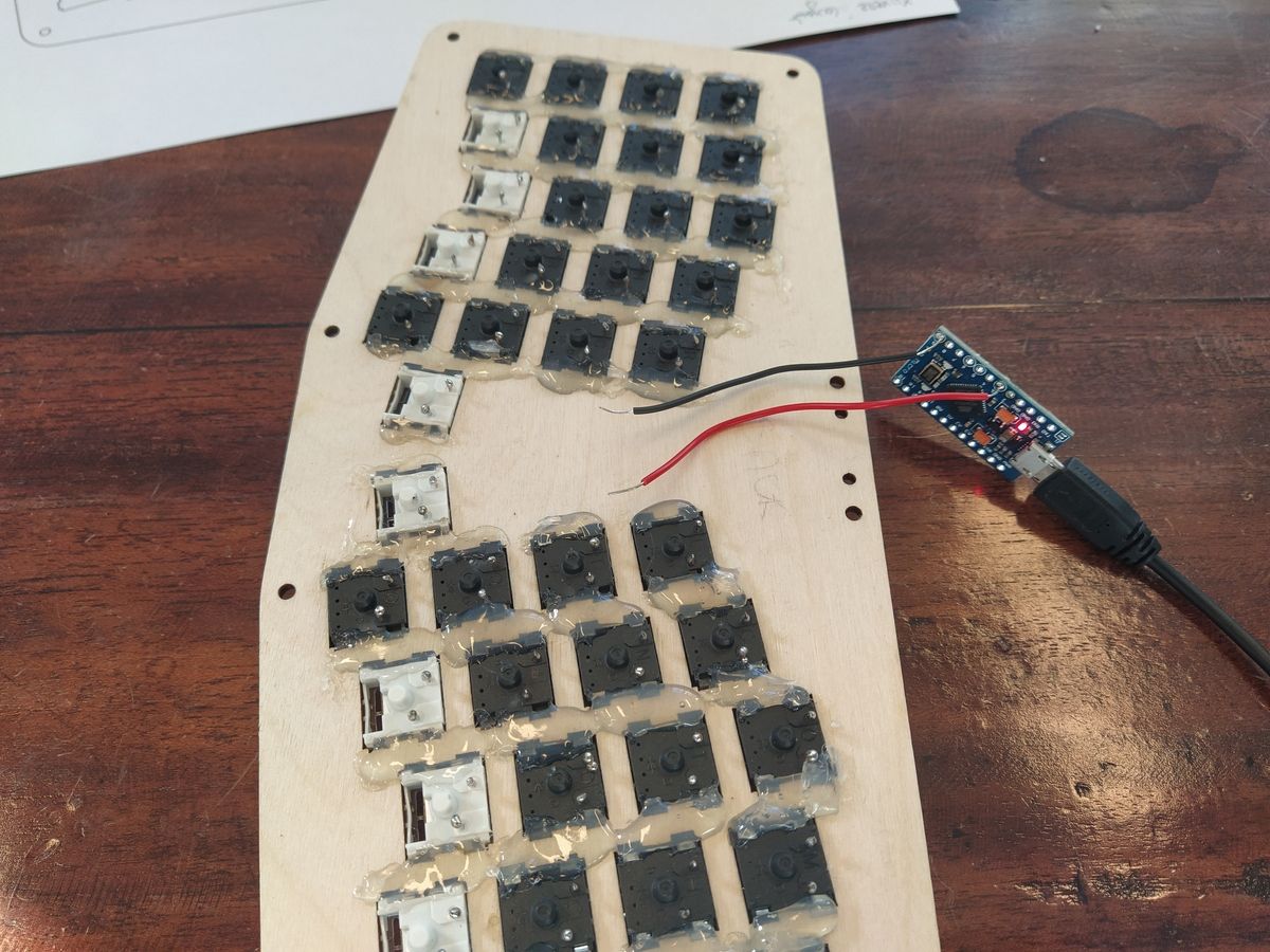 A Pro Micro microcontroller board with red and black test wires touching the two metal pins of a keyboard switch for testing.